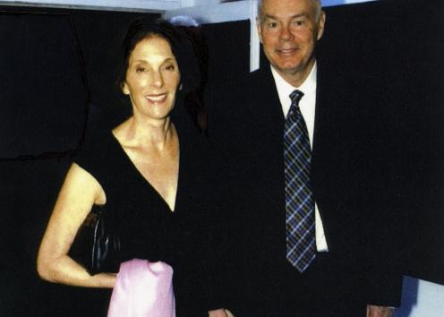 Dr. Braddock in a black suit holding his wife Laura's hand. Laura is holding a pink shawl in her other hand, is wearing a black dress, and is wearing her dark brown hair down.