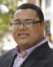 An image of Greg Robinson, a man with short black hair, glasses, and a purple button up shirt and black suit jacket.