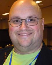 An image of Jason Billehus smiling. Jason is wearing glasses, a green shirt, and a blue lanyard.